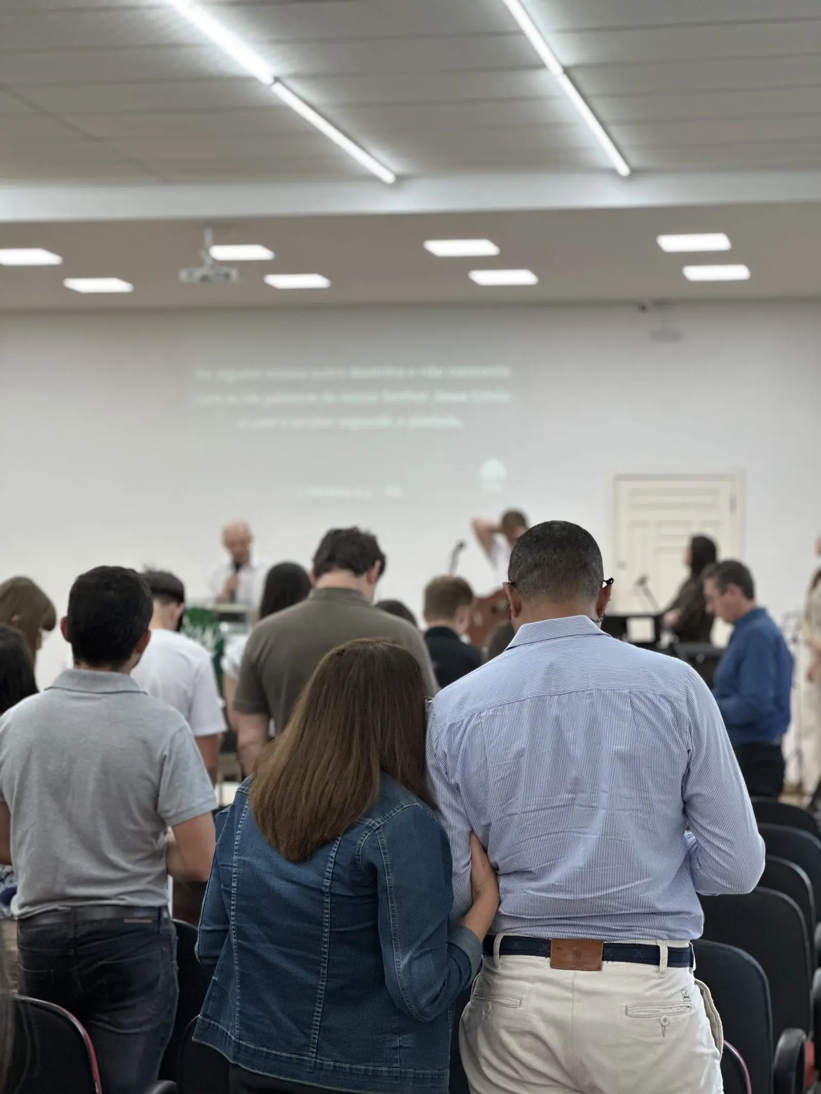 Congregação da IPPB de pé durante o culto, vista de costas, com o pregador à frente e o versículo projetado na parede