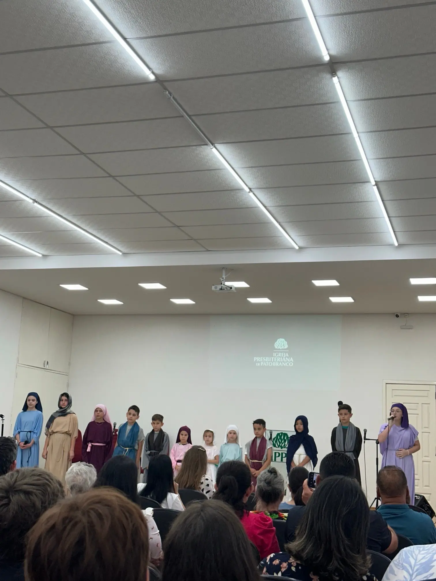 Crianças com trajes bíblicos apresentando uma cantata à frente do salão, diante da congregação