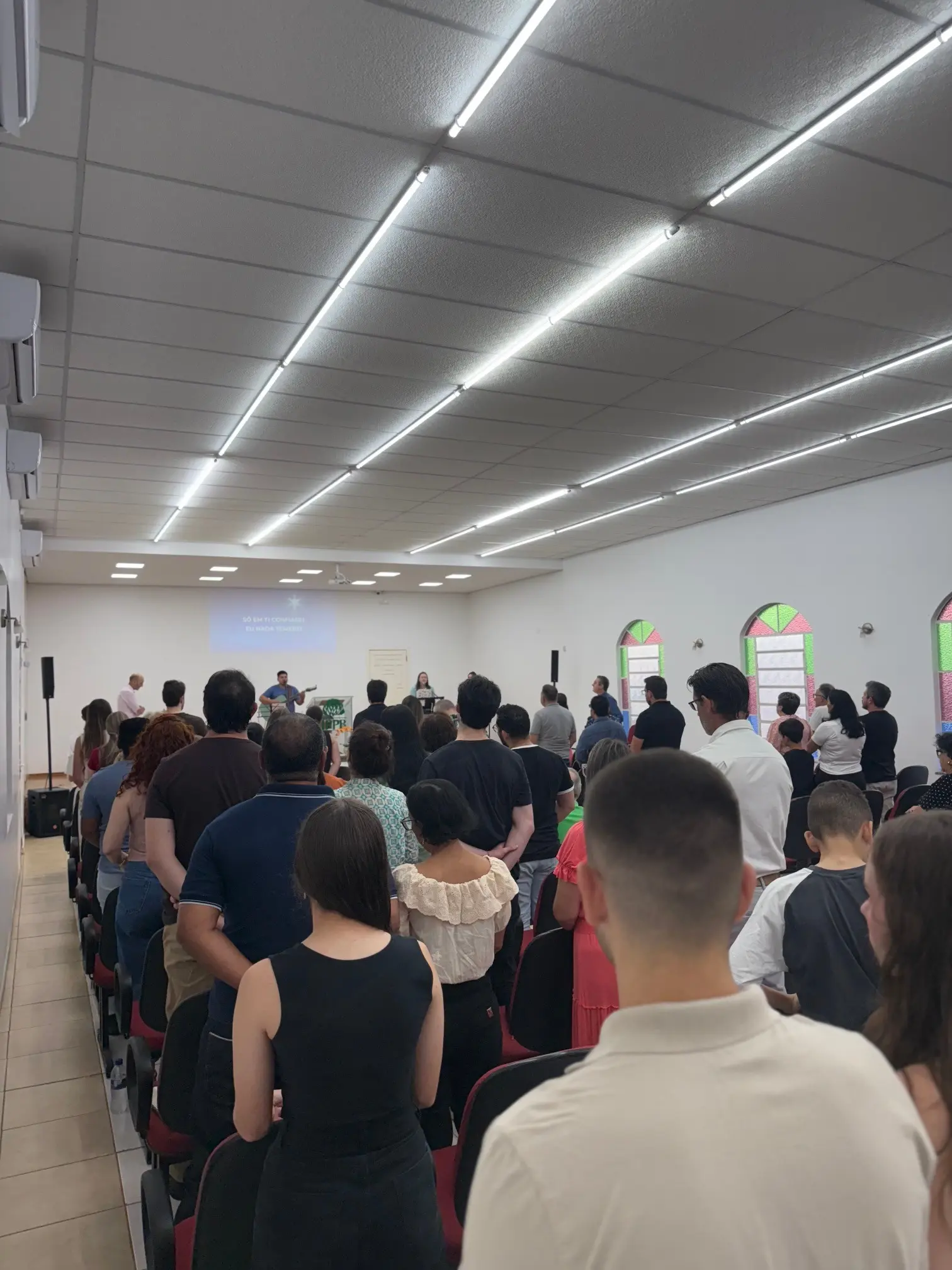Congregação de pé cantando durante o Culto de Adoração, vista do fundo do salão, com a letra do cântico projetada à frente