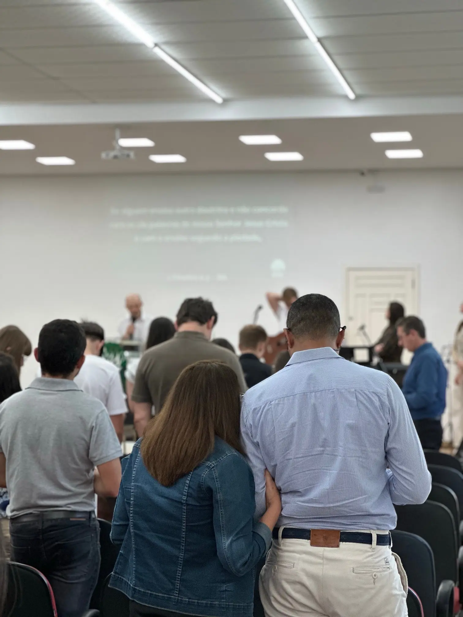 Congregação de pé e de cabeça baixa durante a oração, com um casal de braços dados em primeiro plano