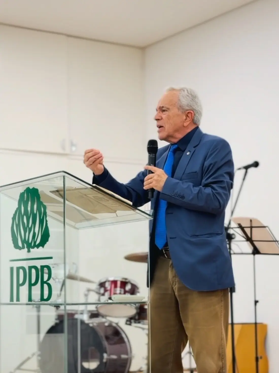 Pregador de paletó azul ao púlpito de vidro com a sarça e a sigla IPPB, falando ao microfone