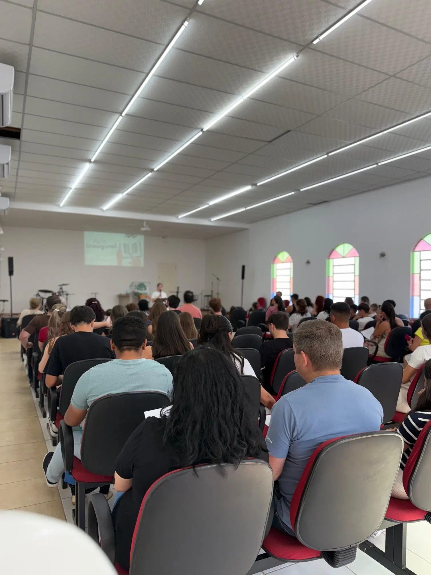 Classe da Escola Dominical reunida no templo, vista do fundo, com a aula inaugural projetada à frente