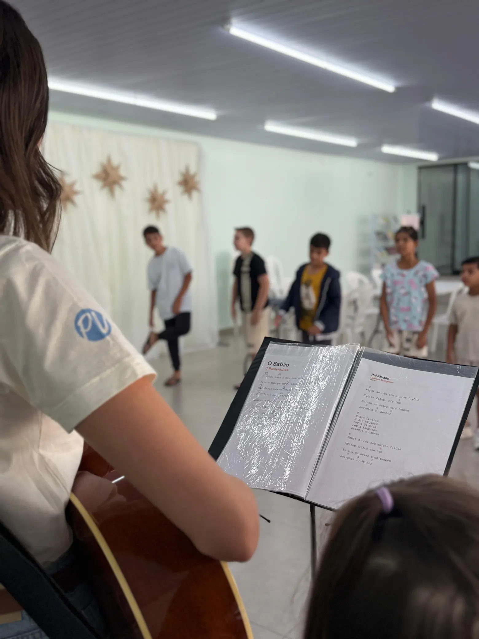 Professora tocando violão enquanto as crianças cantam na classe infantil da Escola Dominical