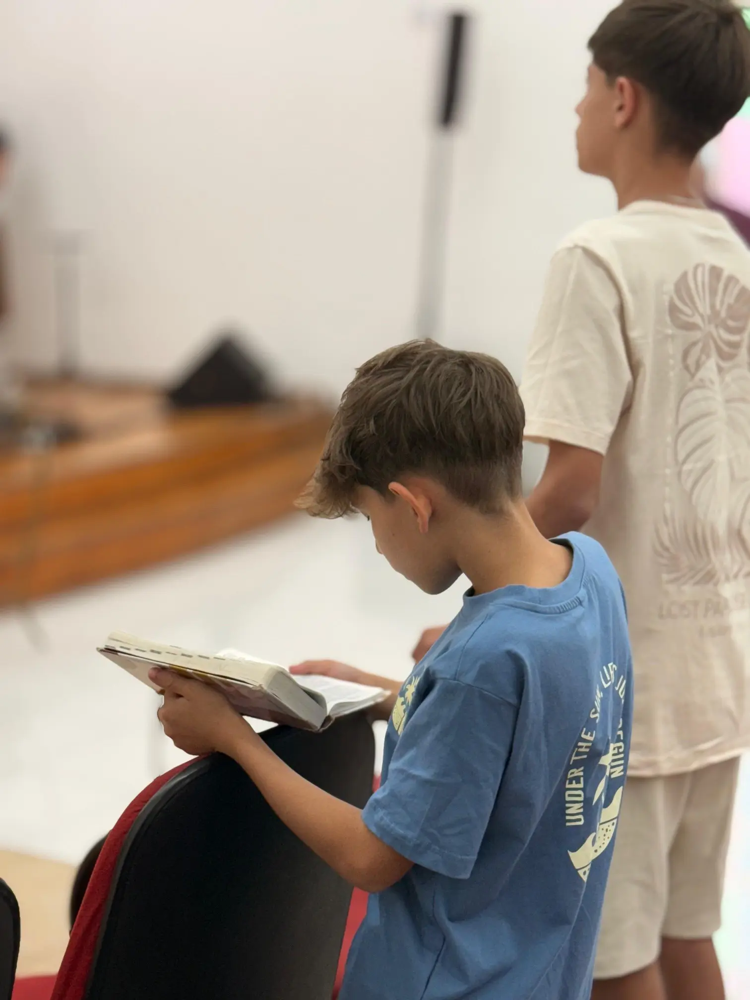 Menino de pé lendo a Bíblia apoiada no encosto da cadeira durante a Escola Dominical