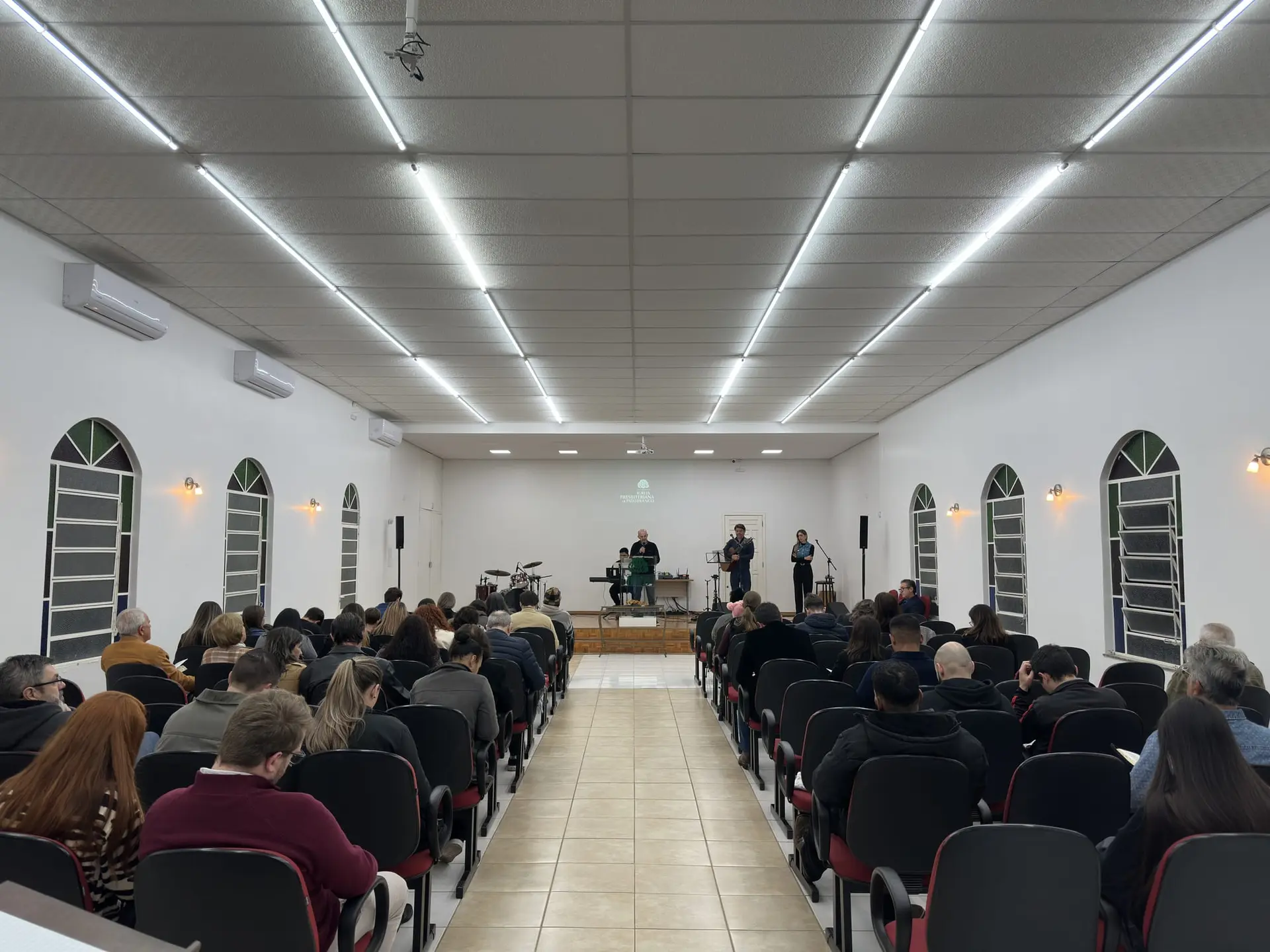 Salão da IPPB cheio durante o culto, visto do fundo, com o corredor central e os músicos à frente