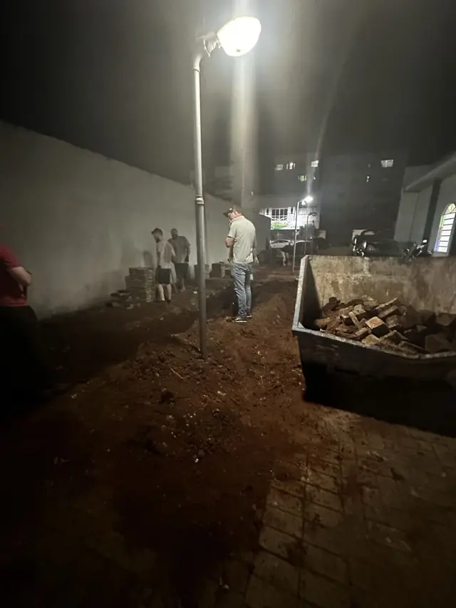 Irmãos trabalhando à noite na obra em frente ao templo, com terra revolvida e uma caçamba de entulho