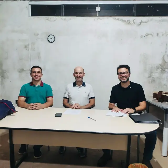 Três homens sorrindo sentados atrás de uma mesa com papéis, numa sala de paredes ainda sem reboco