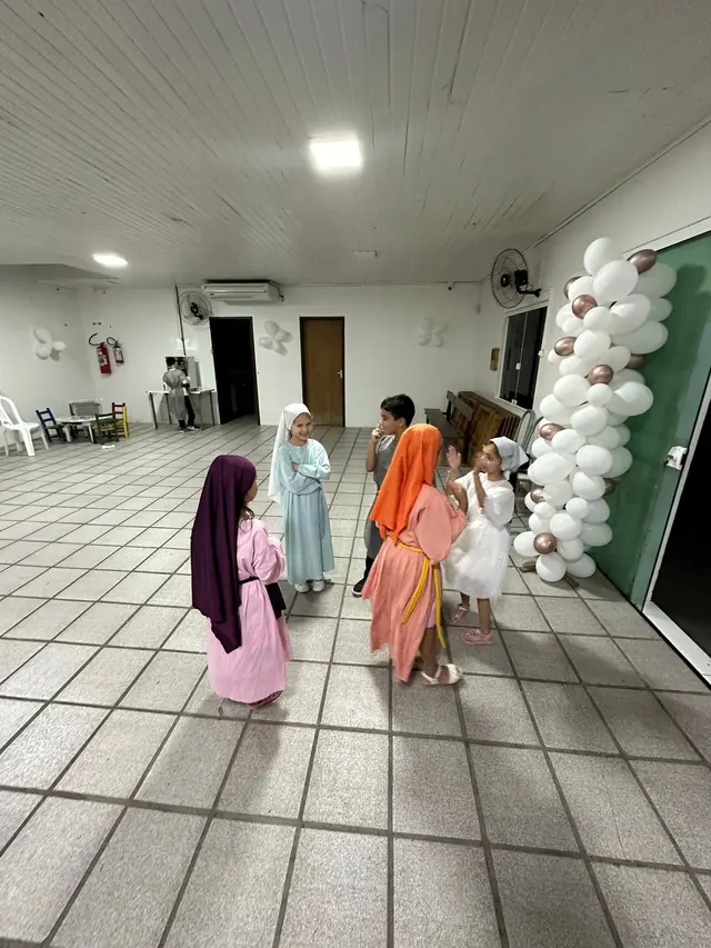 Crianças fantasiadas de personagens bíblicos no salão social, ainda com o piso antigo e o forro de madeira, ao lado de um arco de balões