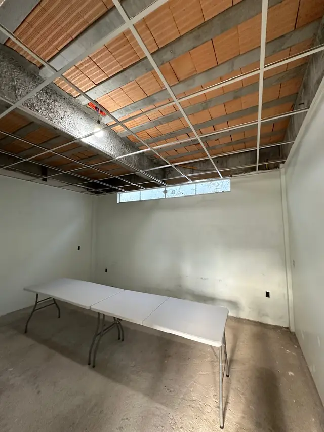A mesma sala ainda em obra, com o forro aberto mostrando as telhas e mesas dobráveis sobre o piso de concreto