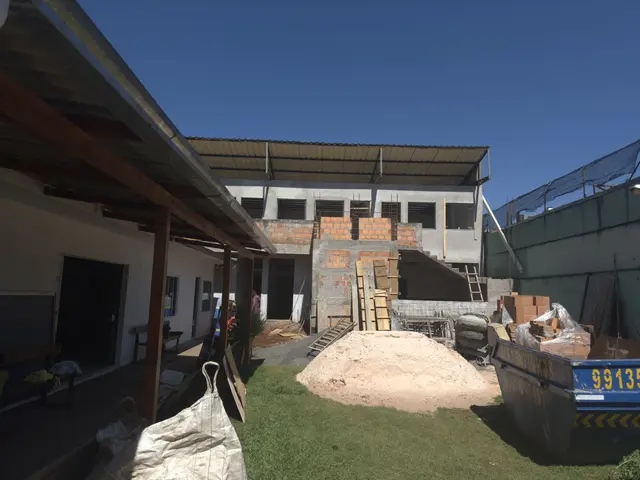Prédio nos fundos do terreno em construção, com tijolos à mostra, areia e caçamba no pátio