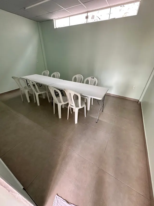 Sala com paredes verde-claras, piso novo e mesas brancas enfileiradas com cadeiras ao redor