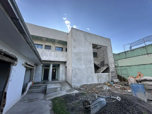Prédio novo no reboco, com a escada de concreto à mostra, entulho e um carrinho de mão no pátio de terra