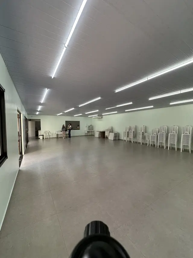 Salão social reformado, amplo, com piso novo, paredes verde-claras, forro branco e luminárias, e cadeiras empilhadas ao fundo