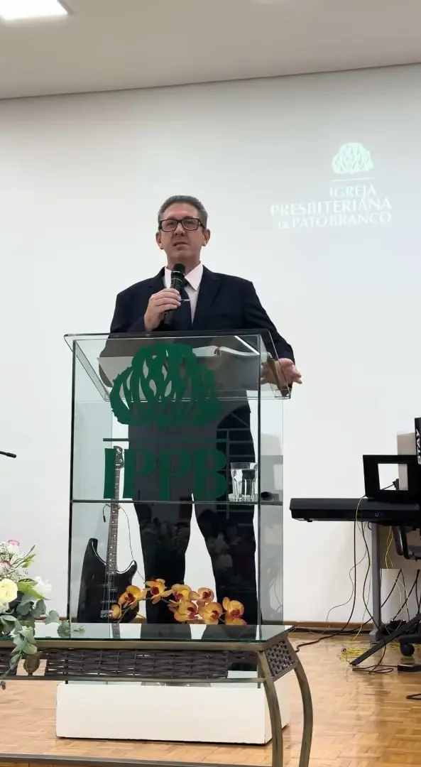 Rev. Maurício, de terno escuro, pregando com o microfone atrás do púlpito de vidro com o logo da IPPB, com o nome da igreja projetado na parede ao fundo
