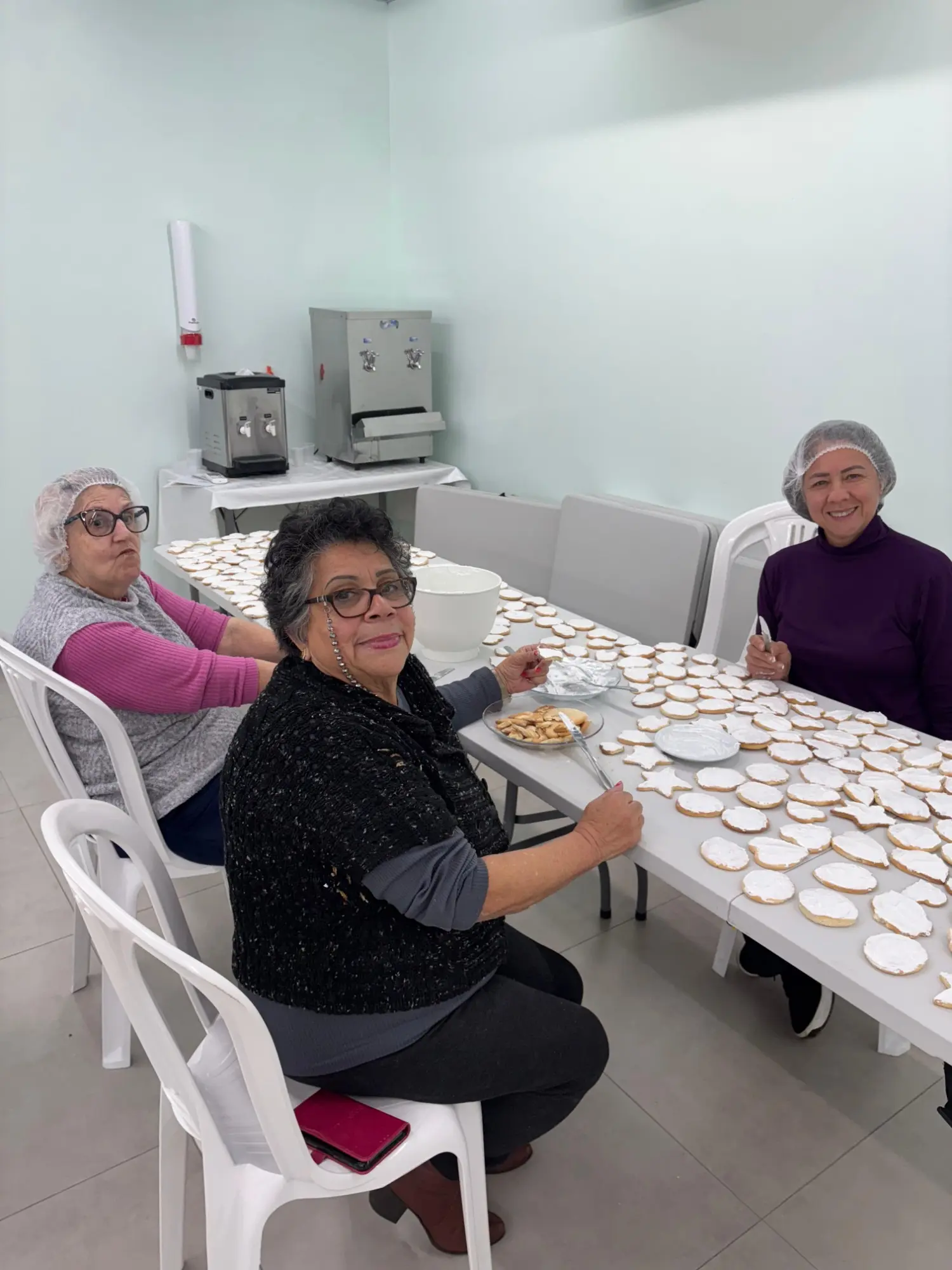 Três mulheres da igreja, de touca, cobrindo bolachas com glacê numa mesa cheia de bolachas