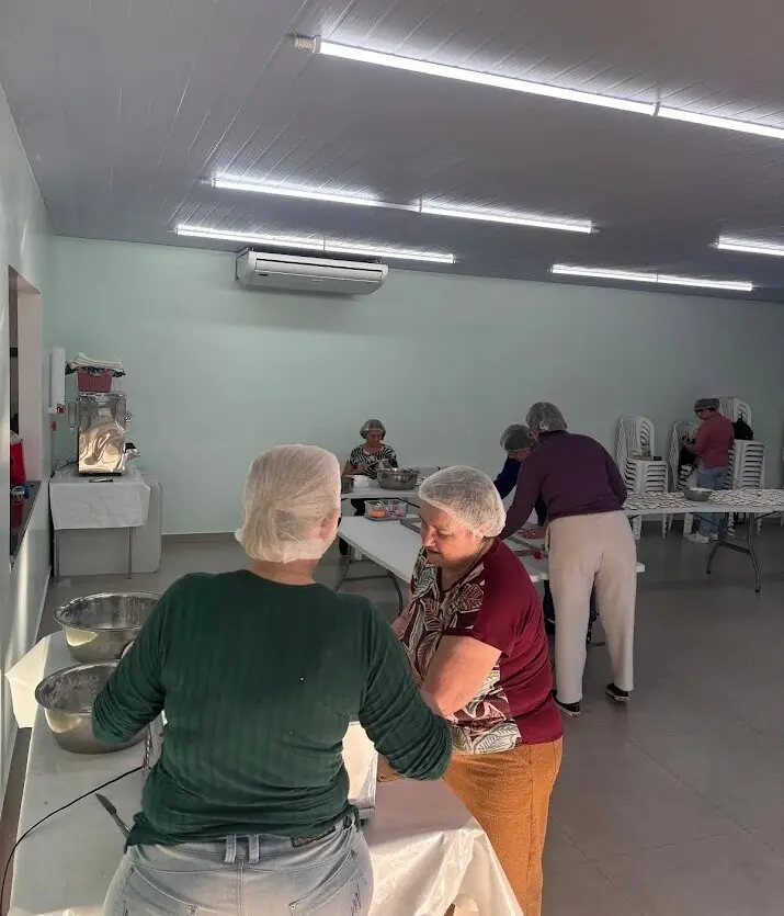Mulheres da igreja, de touca, trabalhando nas mesas do salão da igreja com bacias e utensílios de cozinha