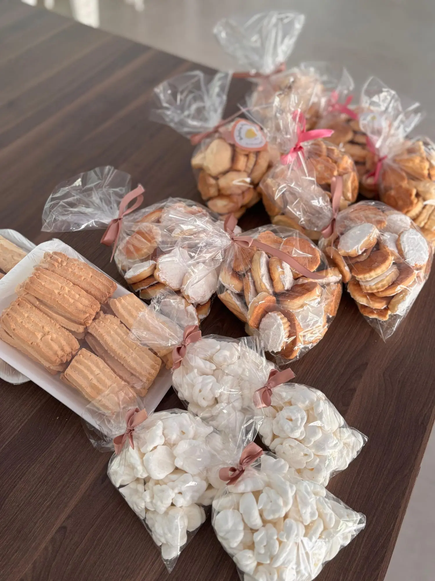 Saquinhos de bolachas, bolachas glaçadas e suspiros do Mãos que Abençoam, amarrados com fita rosa, sobre a mesa ao lado de uma bandeja de biscoitos amanteigados