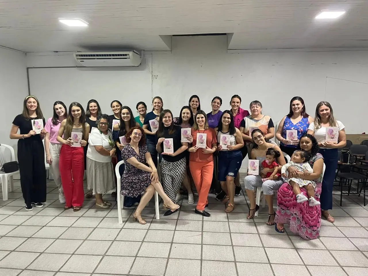 Grupo grande de mulheres do Café Literário, algumas com bebês no colo, mostrando o livro no salão da IPPB