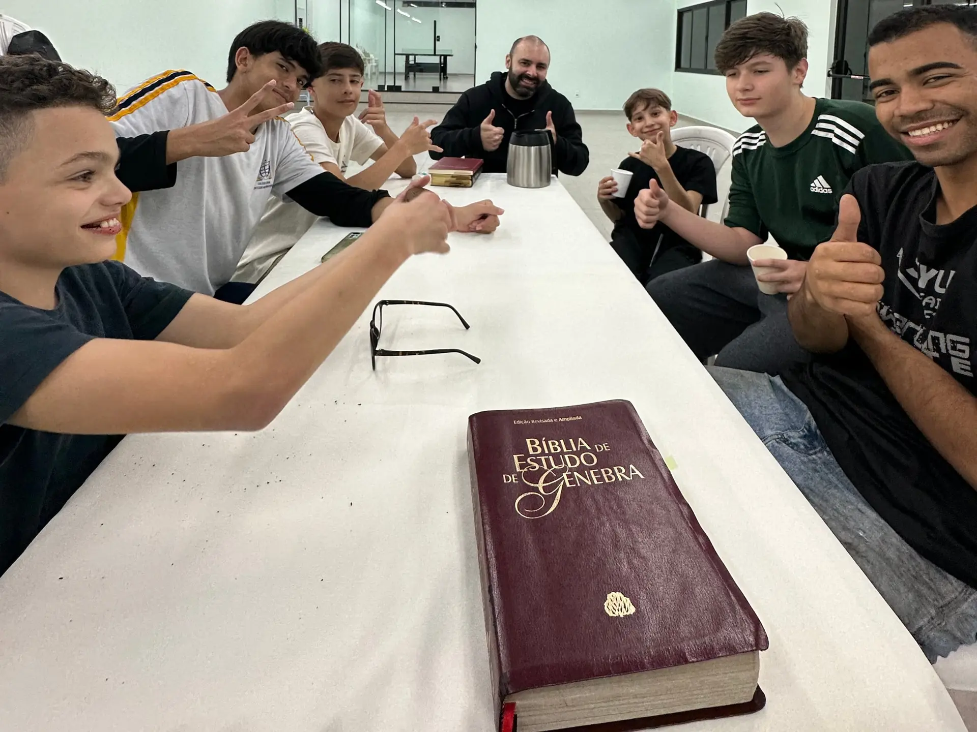 Meninos do Só Meninos e os líderes reunidos em volta de uma mesa com uma Bíblia de Estudo de Genebra, sorrindo e fazendo sinal de positivo