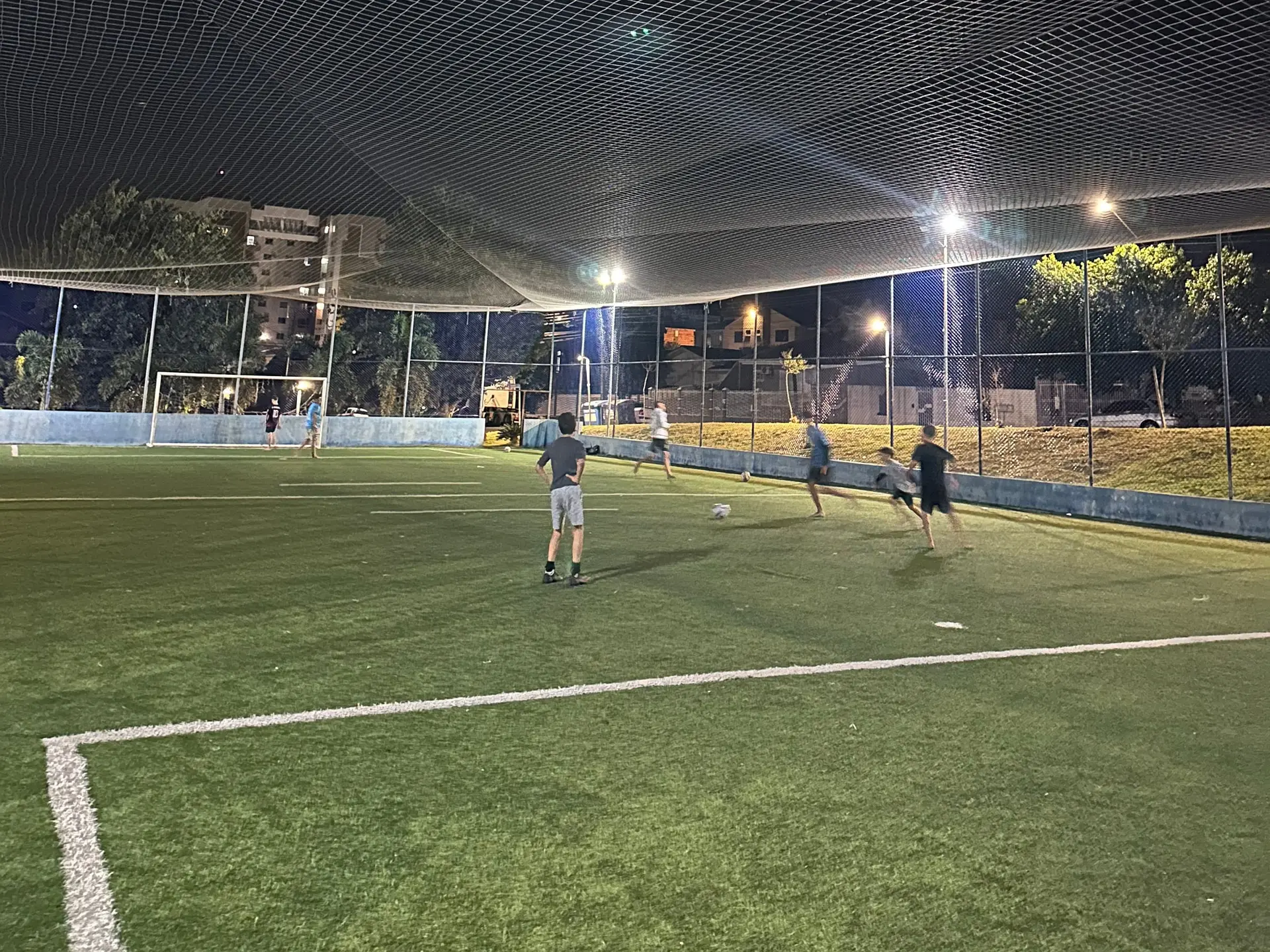 Meninos do Só Meninos jogando futebol à noite numa quadra de grama sintética, vistos de longe