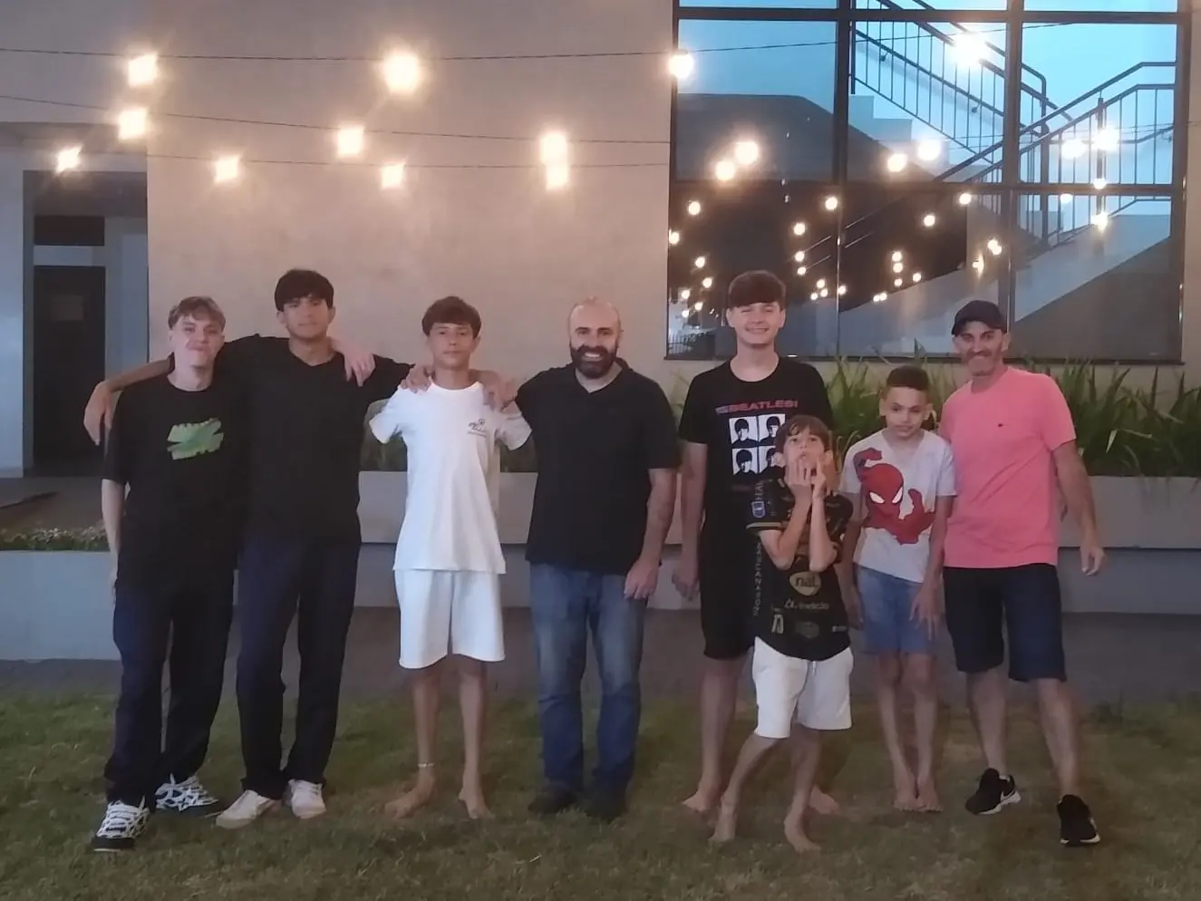 Meninos do Só Meninos e os líderes lado a lado, à noite, no gramado em frente ao prédio da igreja iluminado por varais de luz