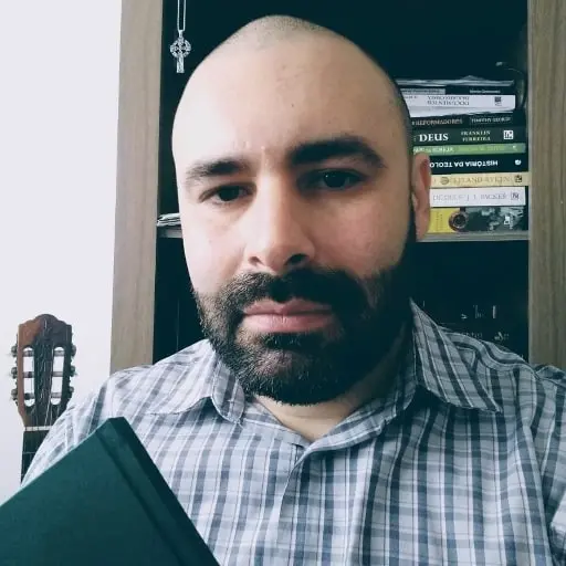 Retrato de Willian Senra, de barba e camisa xadrez, segurando um livro diante de uma estante