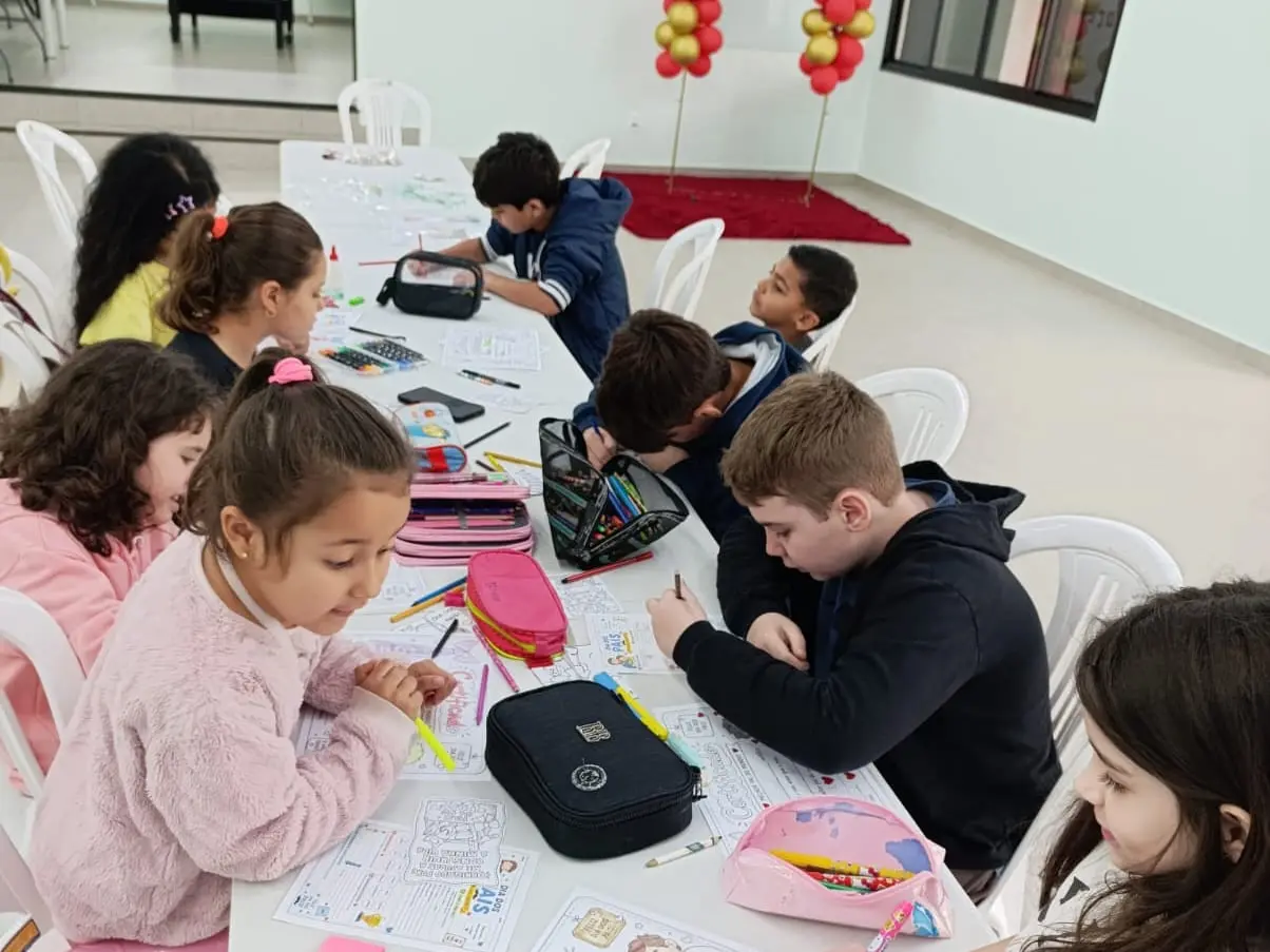 Crianças da UCP sentadas a uma mesa comprida, colorindo atividades do Dia dos Pais com lápis de cor e canetinhas