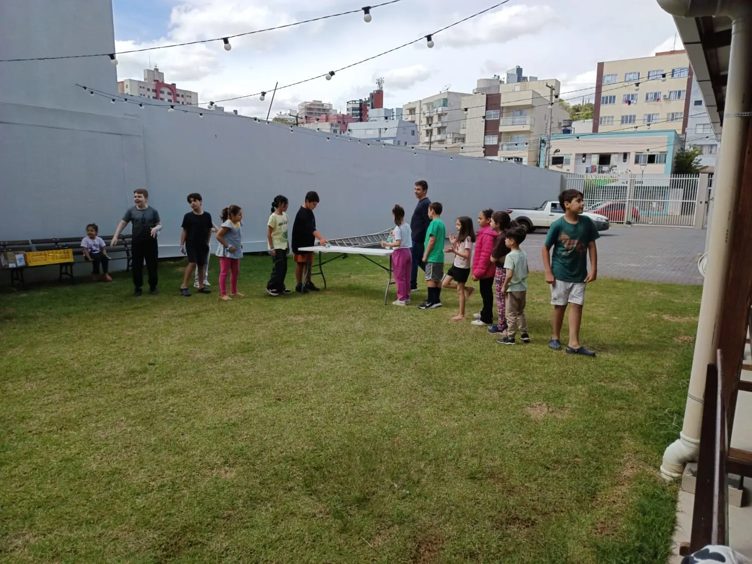 Crianças da UCP em fila no gramado da igreja, numa brincadeira em volta de uma mesa, acompanhadas por um adulto